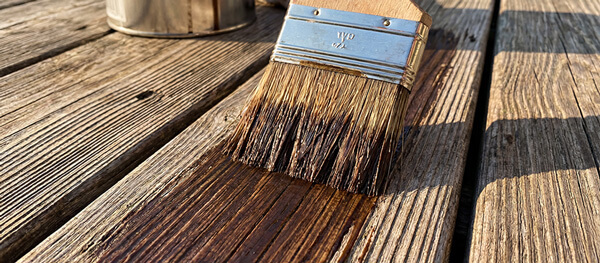 A paintbrush applying a clear wood finish to a wooden surface, revealing the grain. In the background, a paint can is partially visible on a weathered wooden deck.