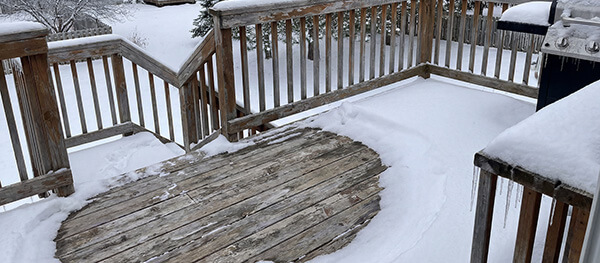 A snow-covered wooden deck with a circular section visible in the center. Snow is piled around the edges and on the railing. A grill is seen in the background, and trees are visible in the distance, creating a winter scene.